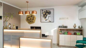 Zona de recepción de un hotel con un mostrador de madera, un letrero en la pared que dice 'hotel ana', una obra de arte enmarcada, un cartel de recepción y una estantería con varios objetos, incluyendo un reloj y un cuenco.
