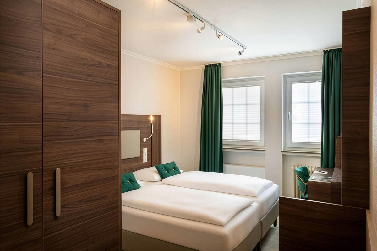 A hotel room with a double bed, wooden wardrobe, a mirror, and a desk with a chair.