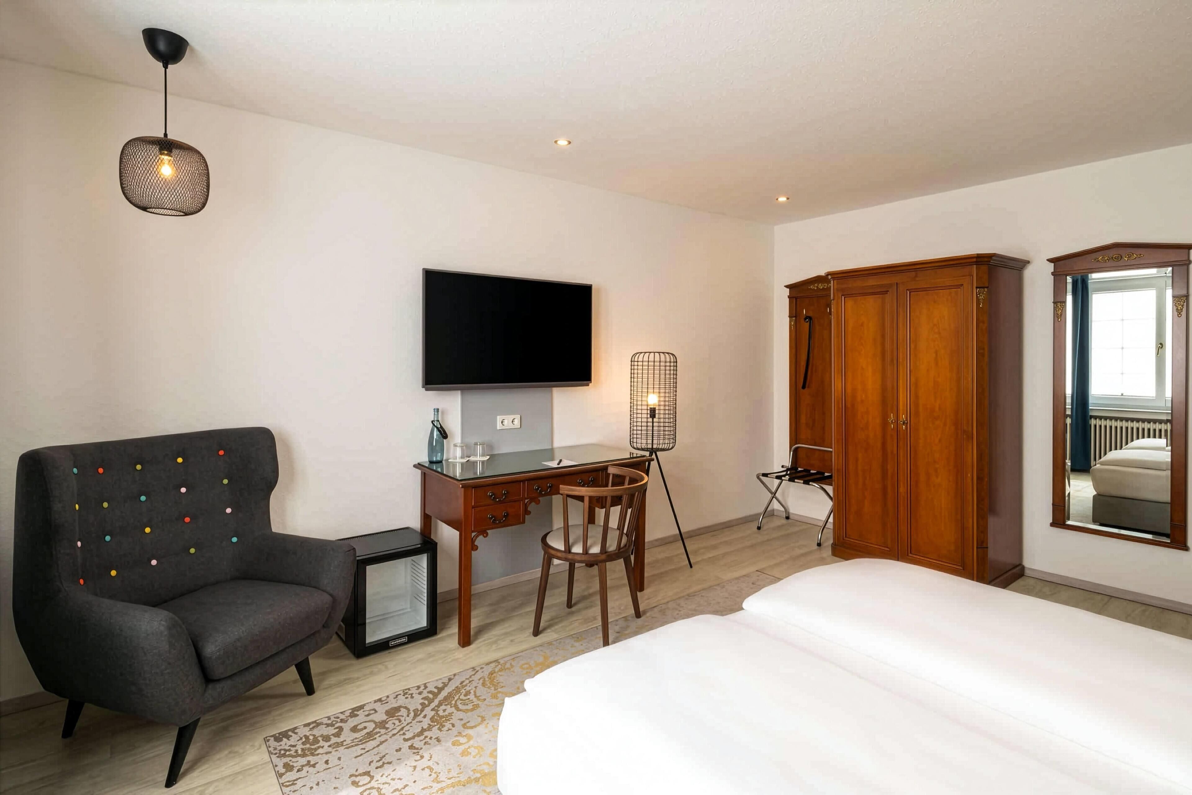 A hotel room with a bed, a desk with a chair, a television, a wardrobe, and a small table with a bottle and glasses.