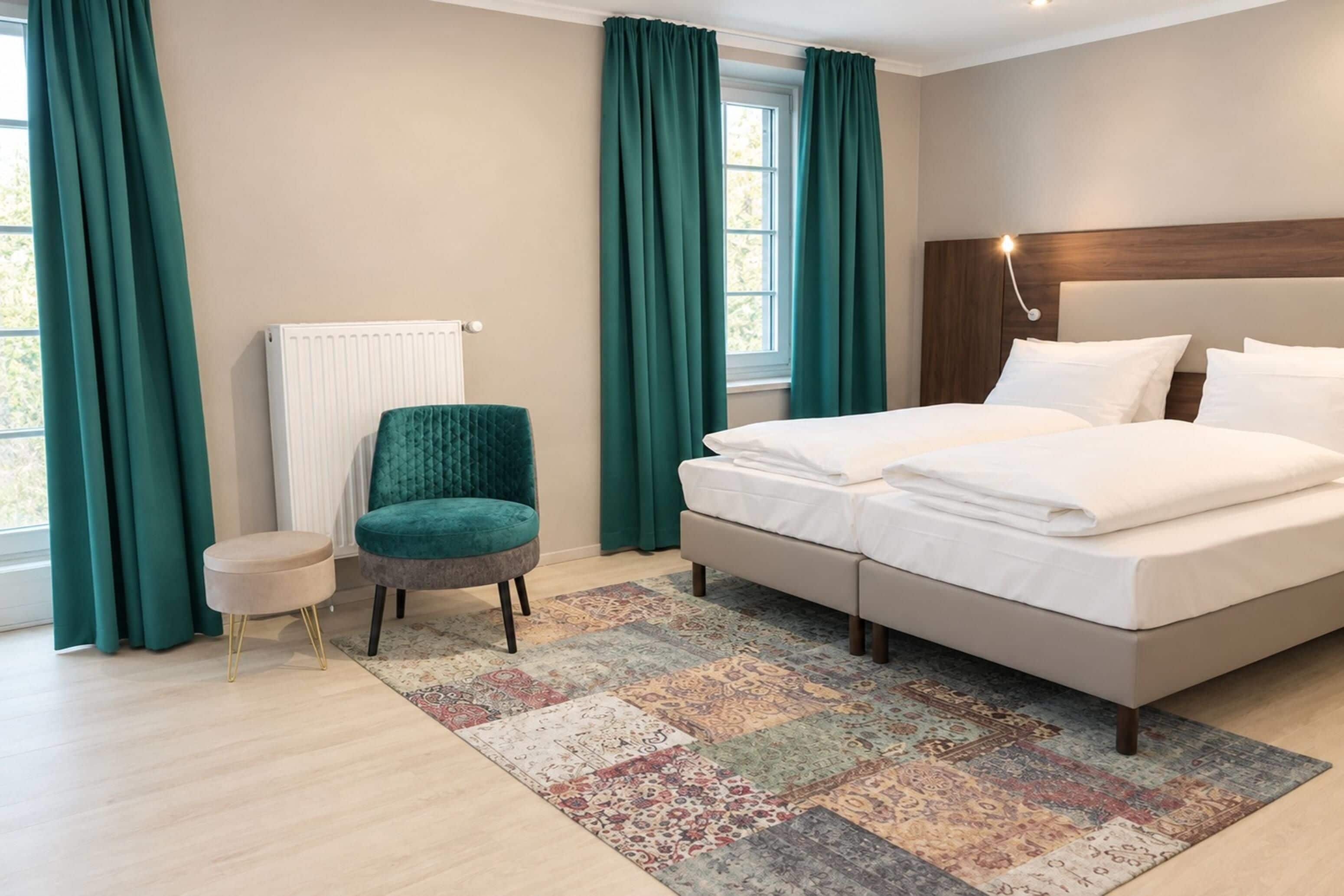 A hotel room with a bed, a chair, a small table, and a window with curtains.