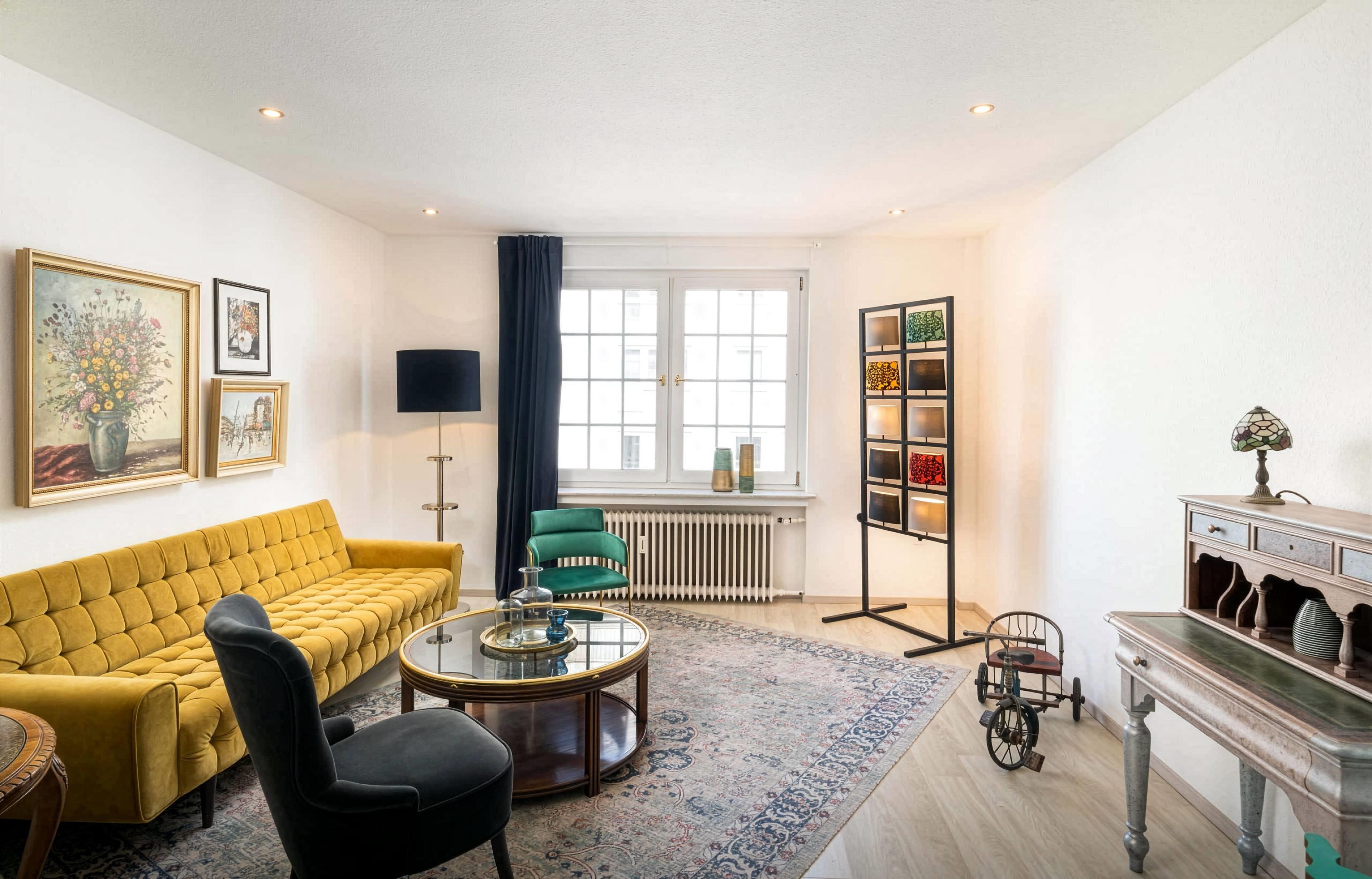 Uma sala de estar com um sofá amarelo, uma poltrona preta, uma mesa de centro de vidro, um poltrona verde, um abajur, um radiador, uma estante e uma luminária decorativa.