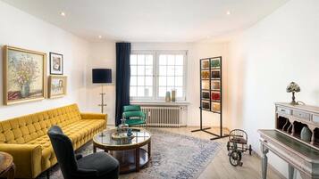 Uma sala de estar com um sofá amarelo, uma poltrona preta, uma mesa de centro de vidro, um poltrona verde, um abajur, um radiador, uma estante e uma luminária decorativa.