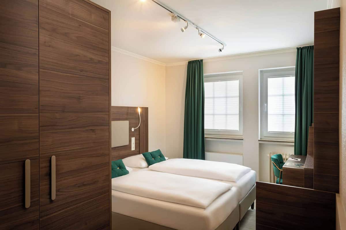 A hotel room with a wooden wardrobe, a bed with white bedding, green curtains, and a desk with a chair.