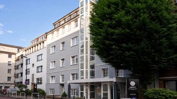 Un hotel de varios pisos con un diseño moderno, que destaca por una entrada con un cartel y un árbol en frente.