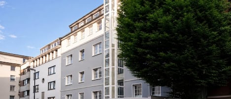 Un hotel de varios pisos con un diseño moderno, que destaca por una entrada con un cartel y un árbol en frente.