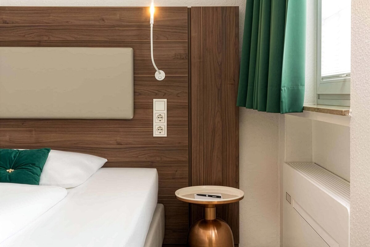A hotel room with a bed, a bedside table with a lamp, a power outlet, and a window with curtains.