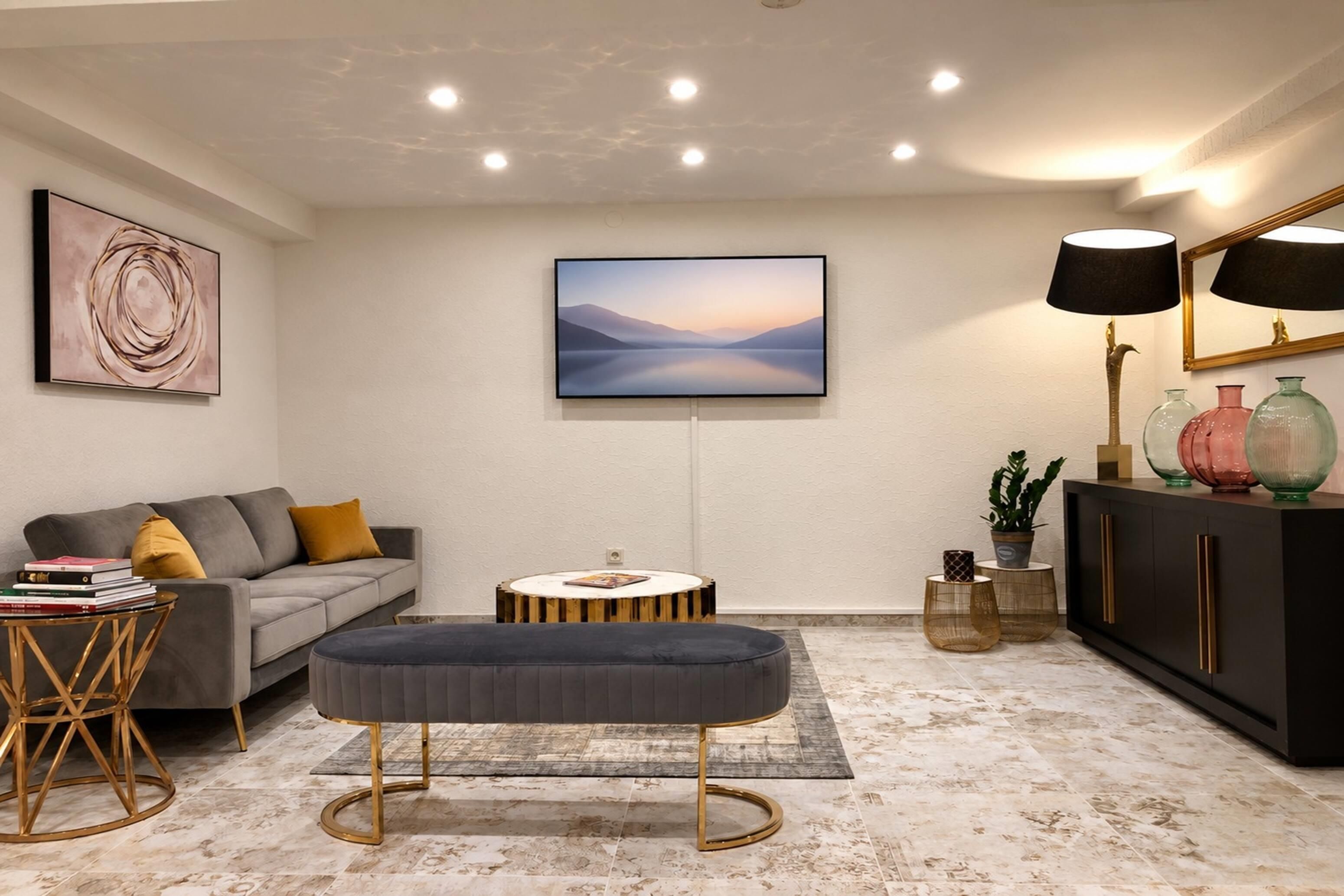 A modern living room with a grey sofa, a round coffee table, and a grey ottoman. There is a painting on the wall, a lamp, and a cabinet with decorative items.