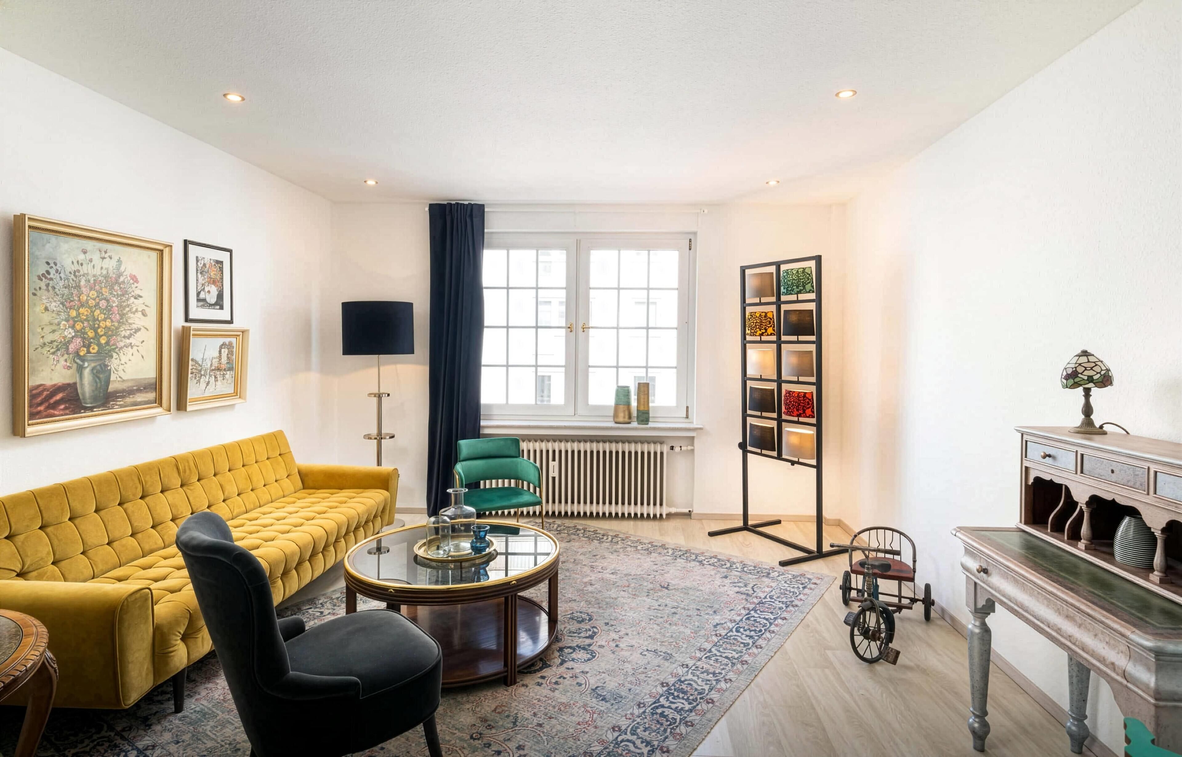 Een woonkamer met een gele bank, een zwarte stoel, een glazen salontafel, een groene fauteuil, een staande lamp, een radiator, een boekenkast en een decoratieve lamp.