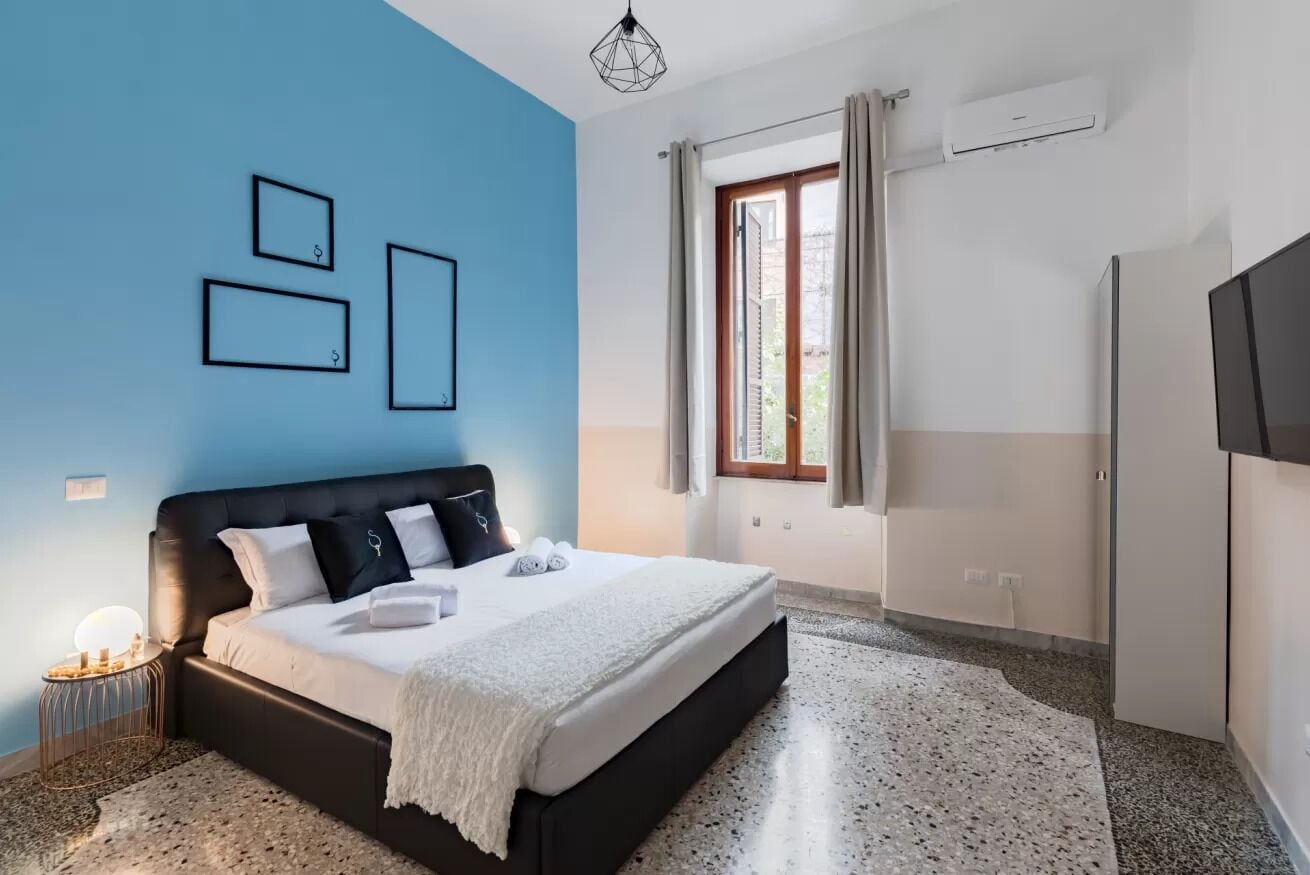 A bedroom with a bed, a window with curtains, a wall-mounted air conditioner, and a TV on the wall.
