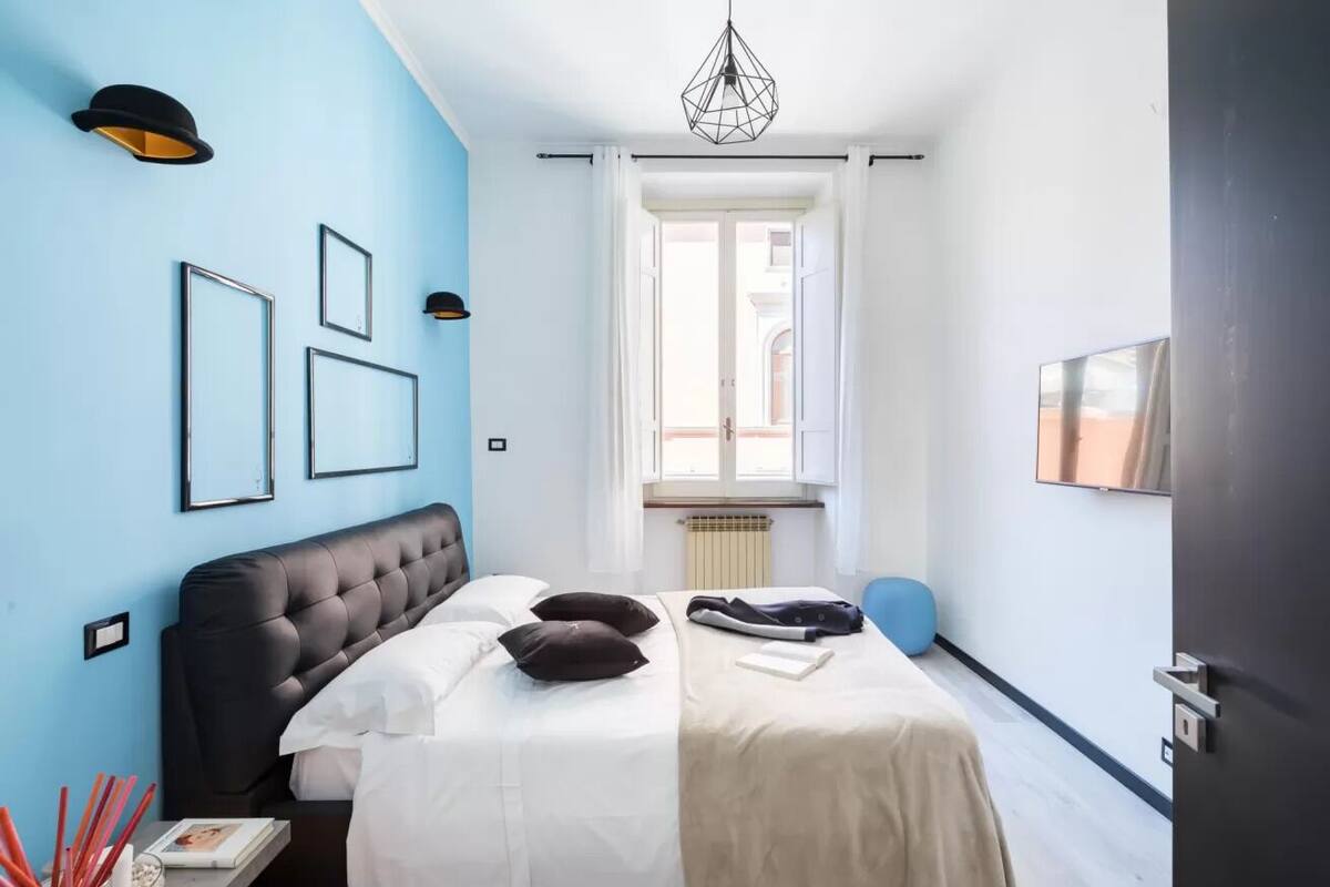 A modern bedroom with a tufted headboard, a bed with white bedding, a blue accent wall, and a large window with curtains.