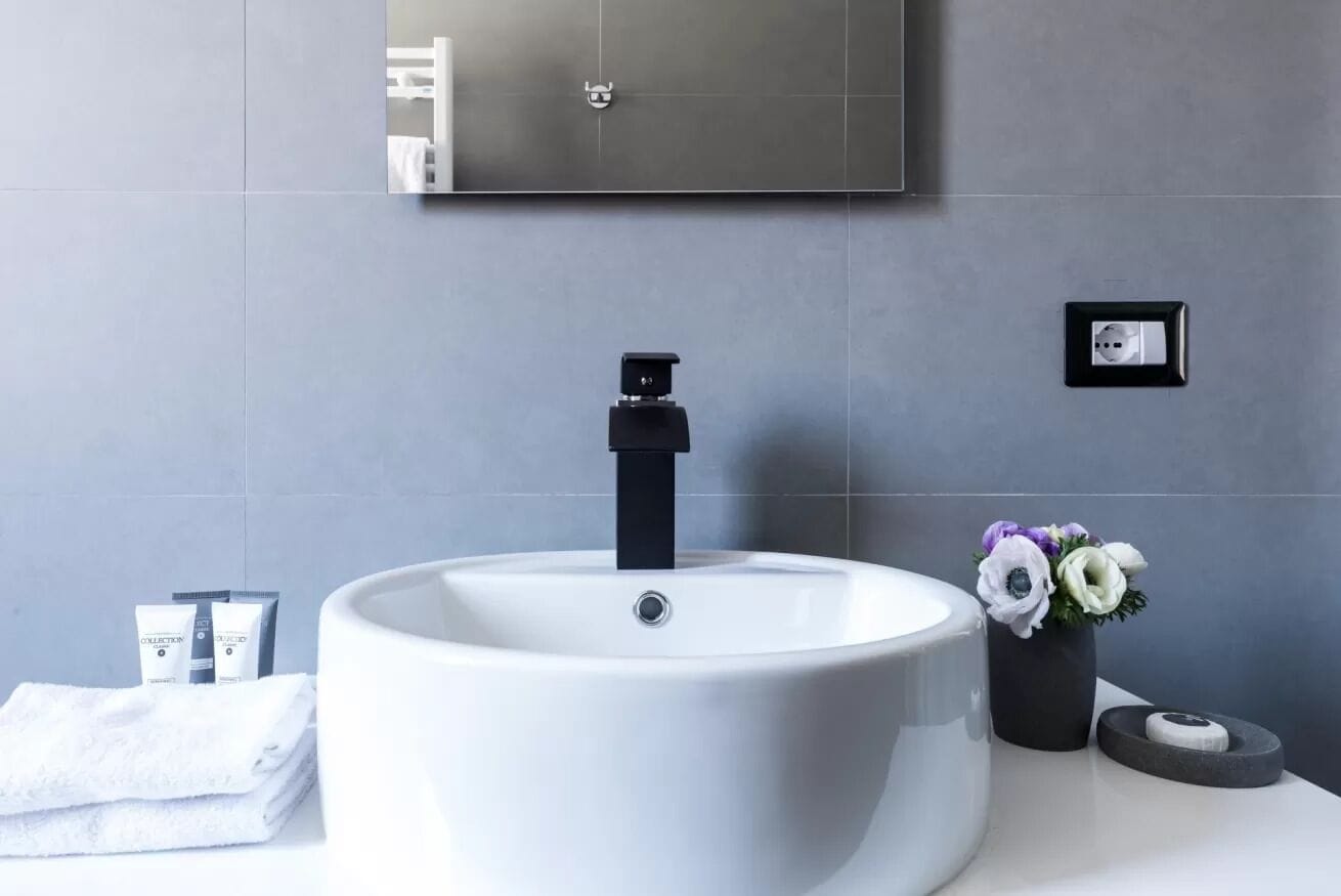 A modern bathroom with a large white sink, a black faucet, a mirror above the sink, and a small black electrical outlet on the wall.