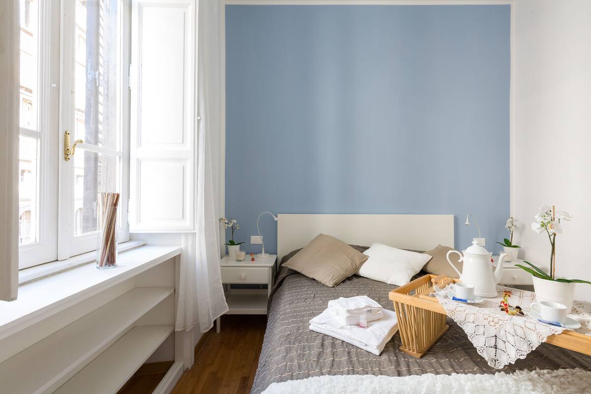 A neatly arranged bedroom with a bed, bedside tables, a window with curtains, and a view of buildings outside.