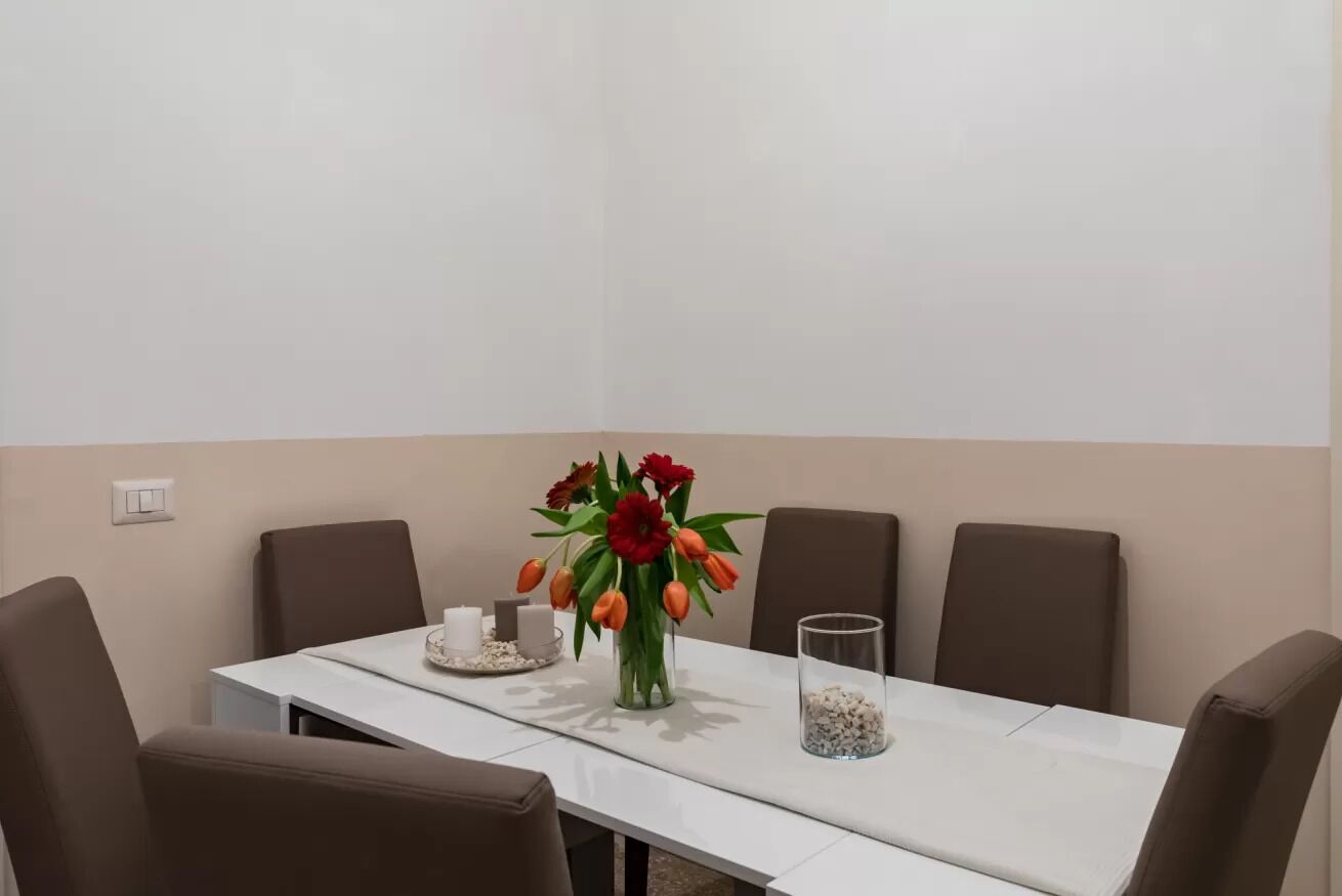 A conference table with four chairs, a vase with red and orange flowers, a glass container with white pebbles, and a small tray with a candle and a cup.