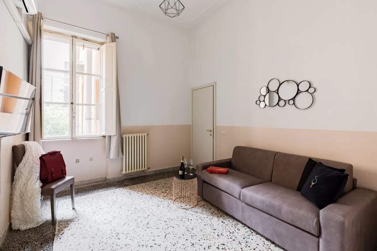 Een moderne woonkamer met een bank, een lange hocker, een klein tafeltje met een fles en glazen, een radiator en een raam met gordijnen.