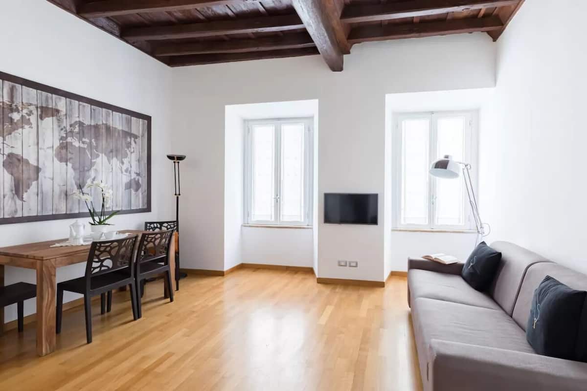 A spacious living room with a wooden floor, a grey sofa, a dining table with chairs, and a world map artwork on the wall.