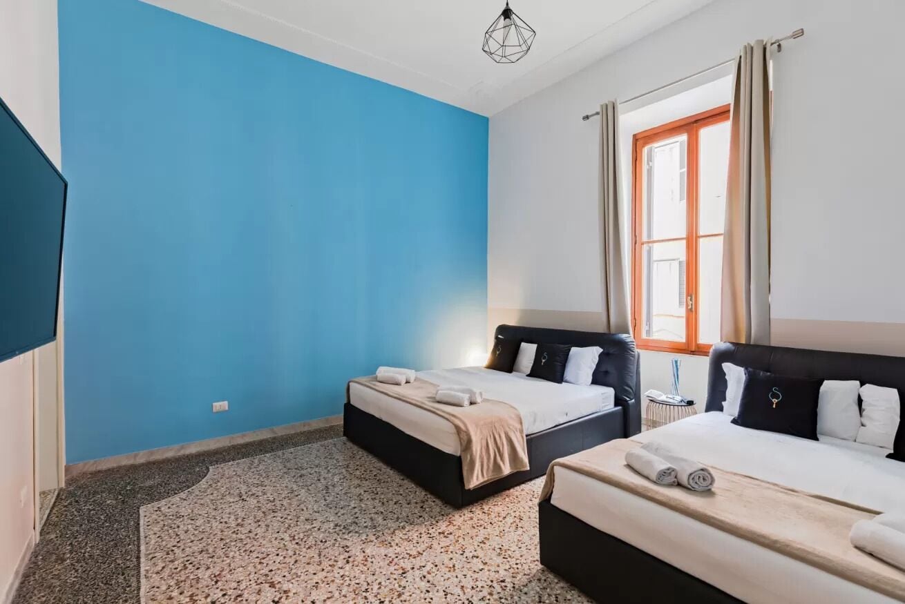 Une chambre avec deux lits, une grande fenêtre avec des rideaux, un luminaire suspendu et une petite table.