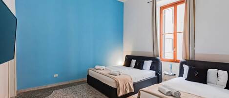 A room with two beds, a large window with curtains, a hanging light fixture, and a small table.