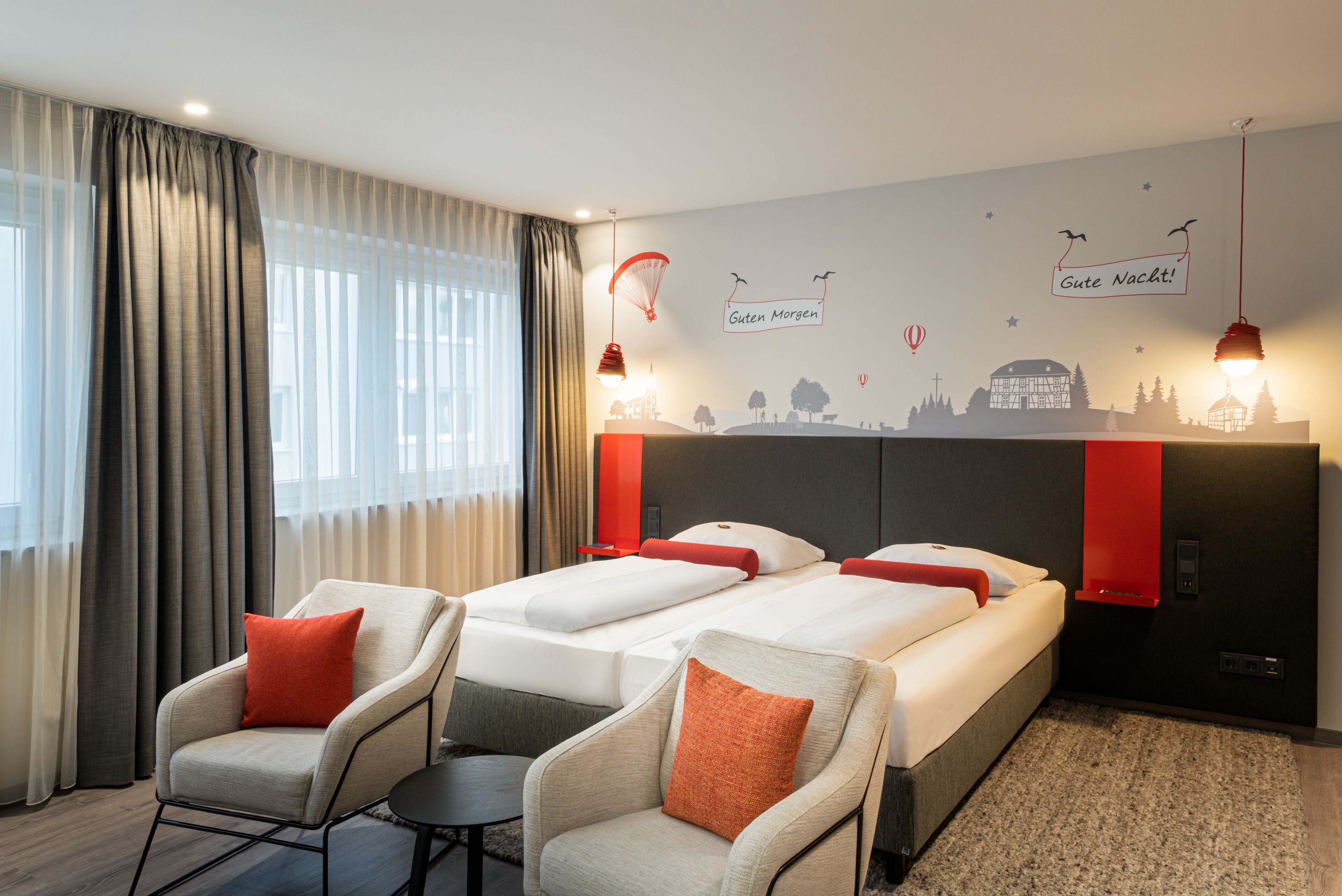 Une chambre d’hôtel avec deux lits, un canapé et un fauteuil. On y trouve des rideaux, une fresque murale et un luminaire au plafond.