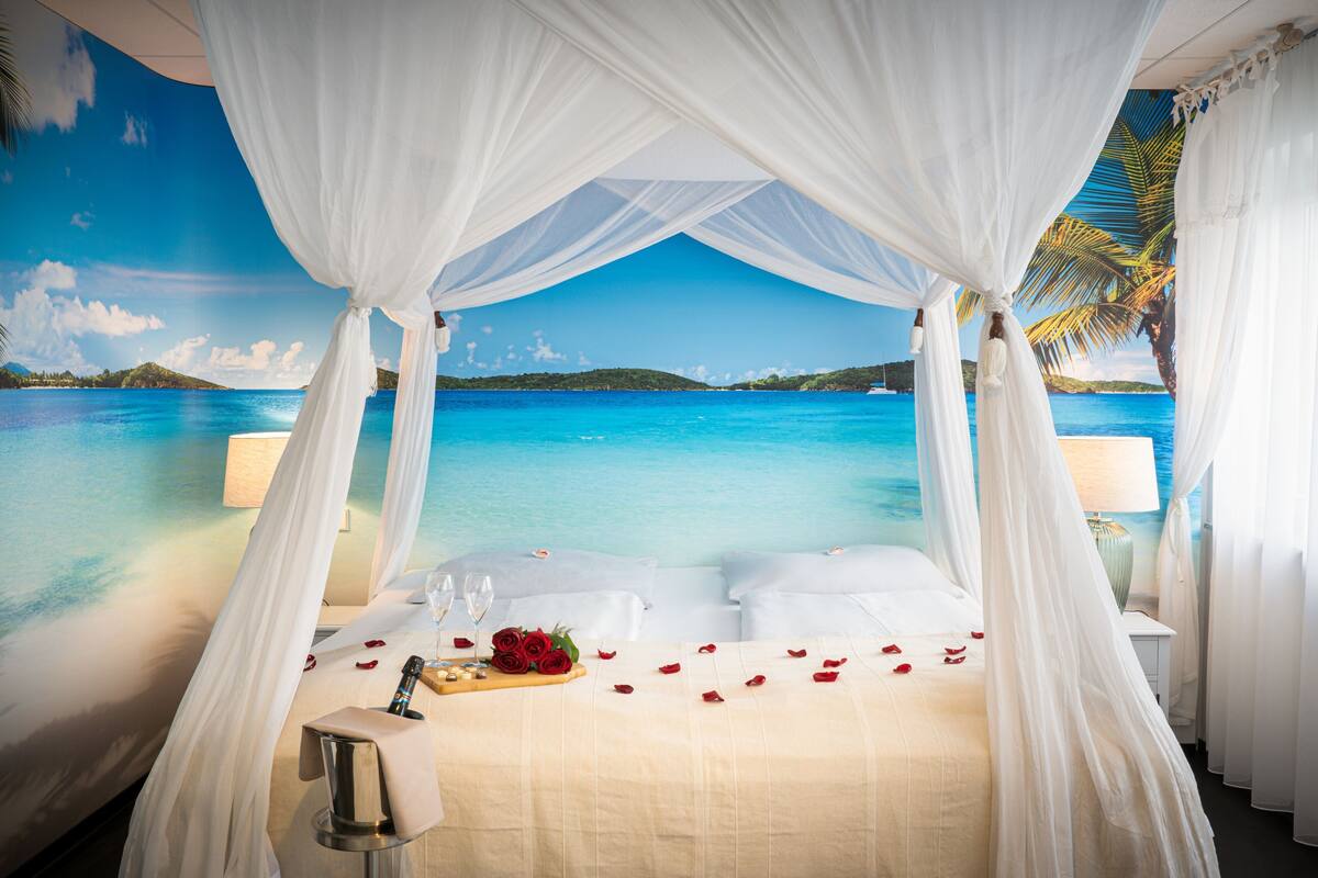 A room with a bed, white curtains, a nightstand, a lamp, and a view of the ocean.