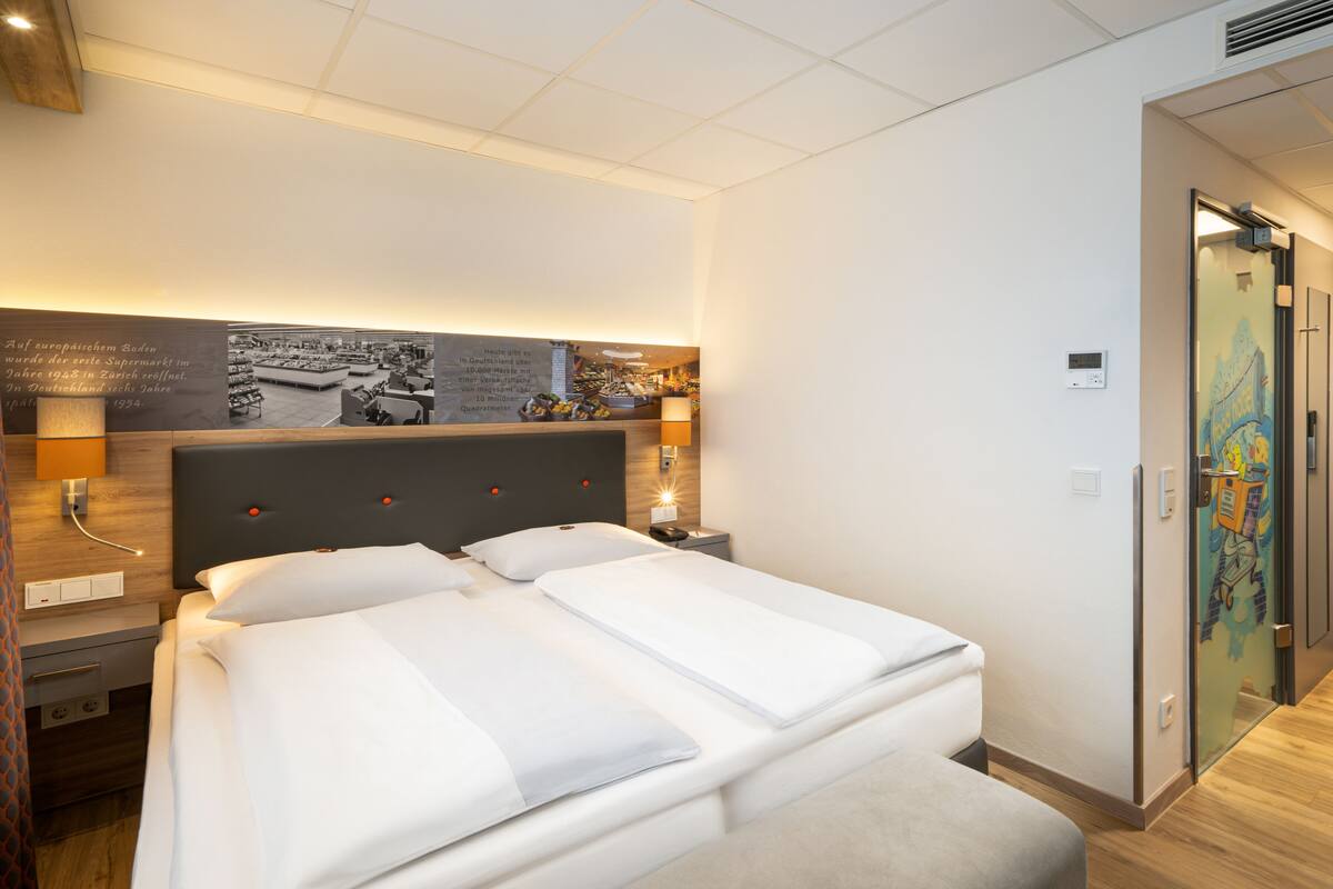 A hotel room with a large bed, a nightstand, a wall-mounted lamp, and a poster.