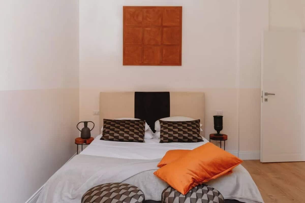 A neatly made bed with patterned pillows, a wooden artwork above it, and a small table with a vase on the side.