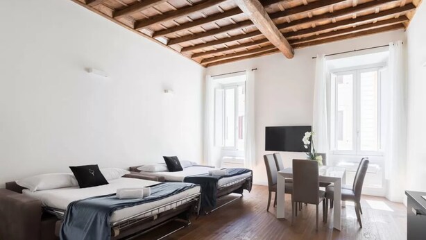 A modern, well-lit room with a wooden ceiling, two beds, a dining table with chairs, and large windows.