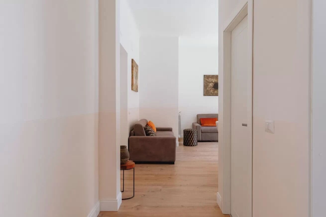 Un pasillo que conduce a una sala de estar con un sofá, una silla, una pequeña mesa con un jarrón y un cuadro en la pared.