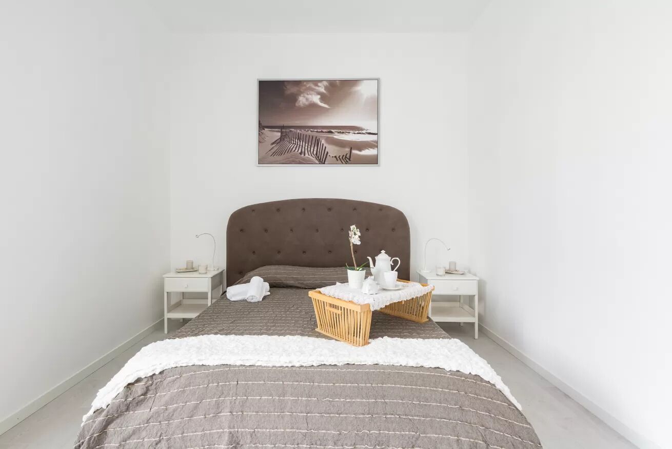 A bedroom with a large bed, a headboard, two bedside tables, a nightstand, a framed picture on the wall, and a small table with a vase and teapot.