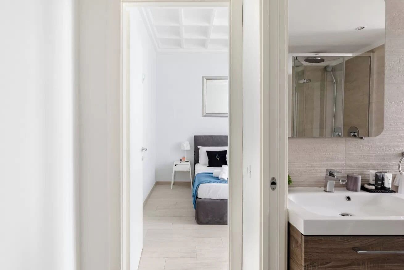 A modern hotel room with a large bed, a bedside table with a lamp, a bathroom with a sink and mirror, and a walk-in shower.
