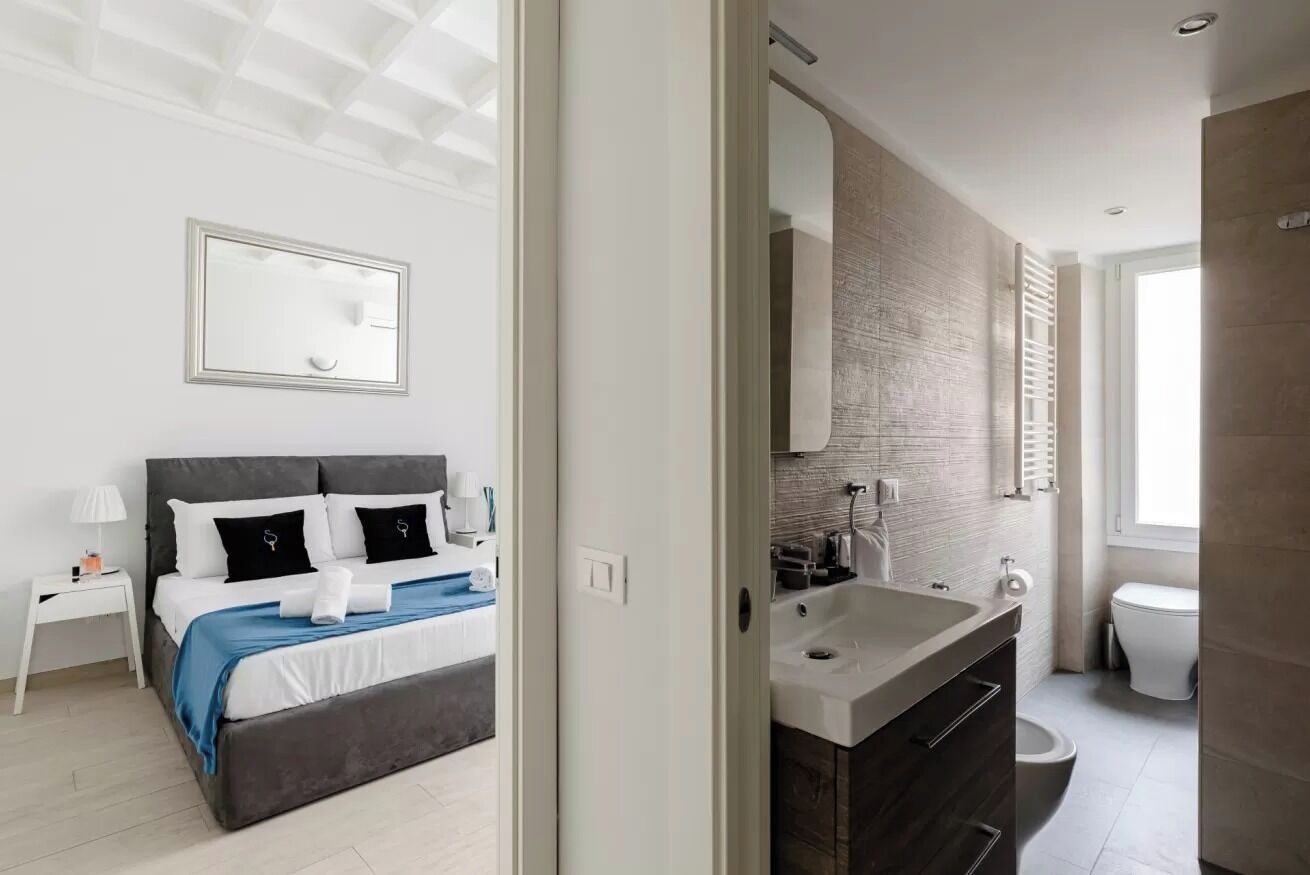 Quarto moderno com uma cama grande, mesas de cabeceira, um espelho e um banheiro com pia e vaso sanitário.