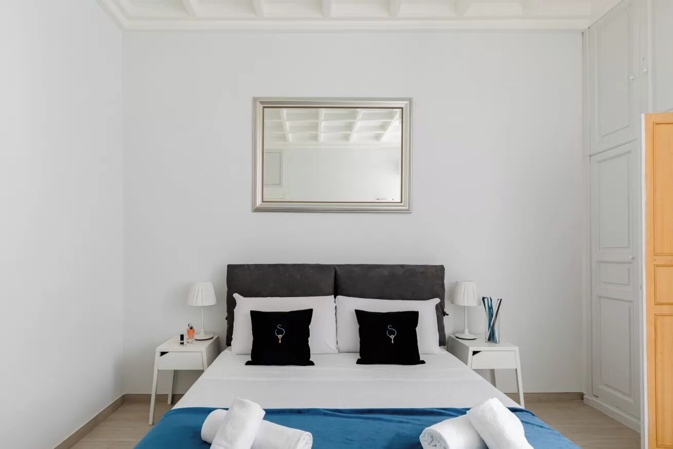 A neatly arranged bedroom with a bed, two bedside tables, a nightstand, a mirror, and a wardrobe.