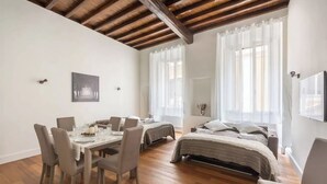 A room with a wooden ceiling, a dining table with chairs, a bed with a headboard, and a window with sheer curtains.