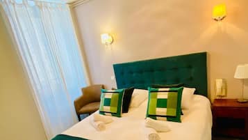 Habitación de hotel con una cama grande, cabecera verde y dos almohadas. Se observan dos apliques de pared y una ventana con cortinas traslúcidas.