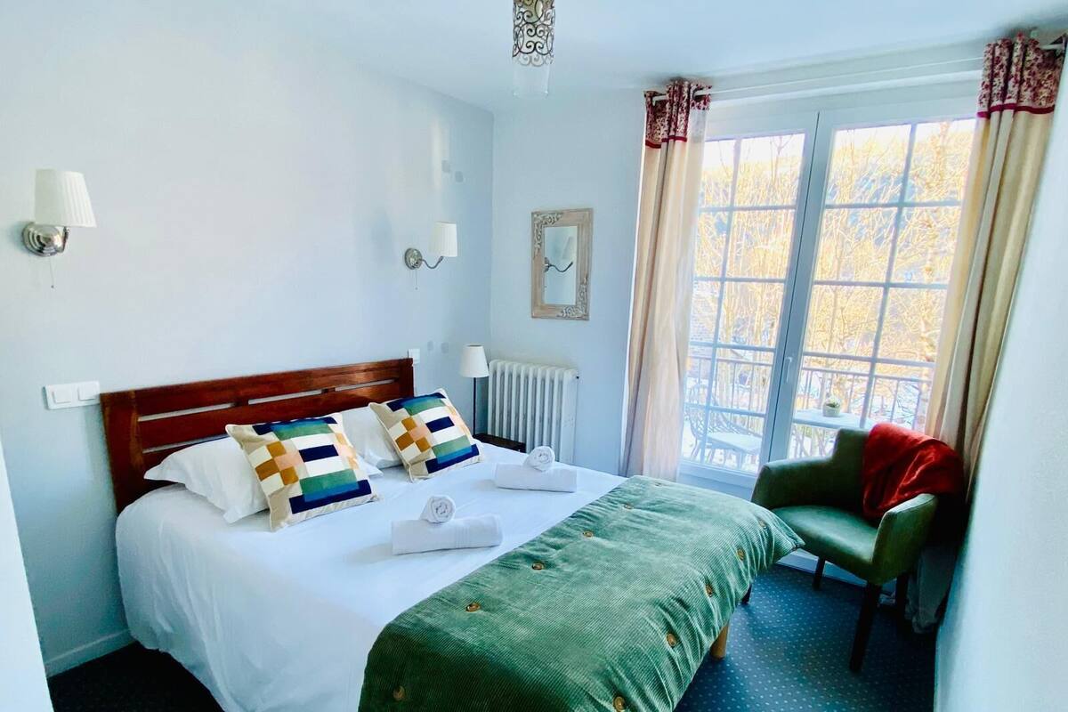 A hotel room with a bed, a green armchair, a window with curtains, and a radiator.