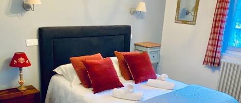 Habitación de hotel con una cama grande, dos lámparas de noche, un candelabro, una ventana con cortinas y una pequeña mesa con lámpara.