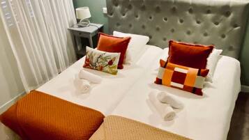 Una cama bien hecha con varias almohadas y una colcha, una mesita de noche con una lámpara y una ventana con cortinas.