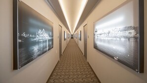 Pasillo de hotel con alfombra estampada, fotografías enmarcadas de un paisaje urbano en las paredes y una lámpara en el techo.