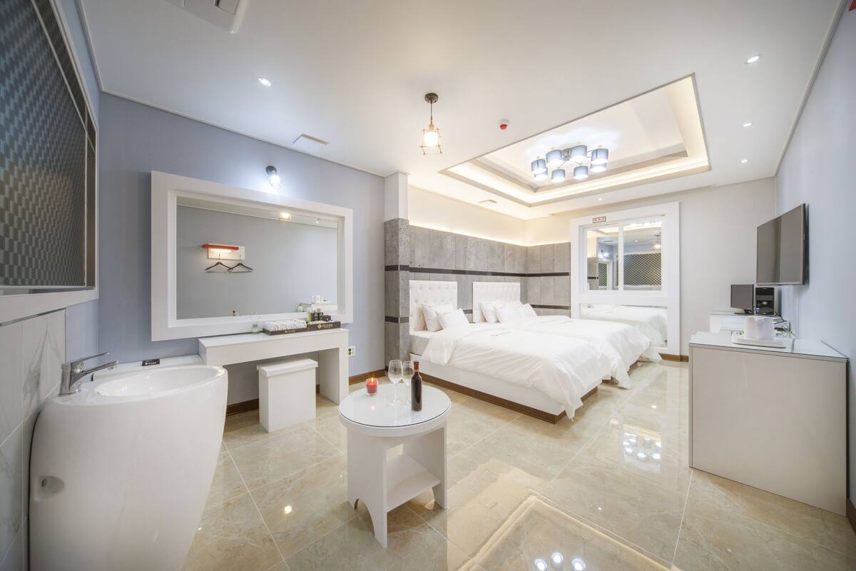 A modern hotel room with a large bed, a sitting area with a table, a vanity with a mirror, and a large window.