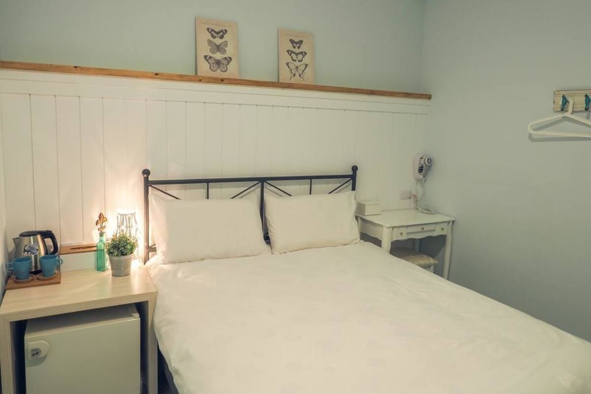Ein Schlafzimmer mit Bett, Nachttisch und kleinem Schreibtisch. Über dem Bett befinden sich zwei gerahmte Schmetterlingsbilder an der Wand.