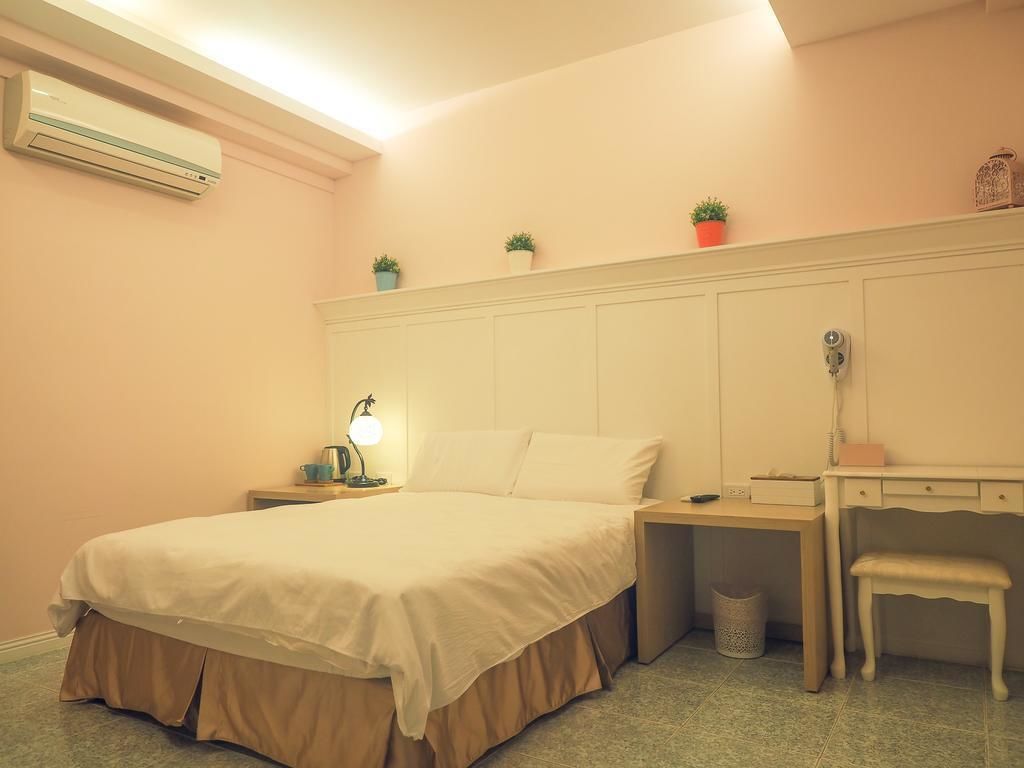 A bedroom with a bed, bedside table, lamp, desk, chair, and potted plants.