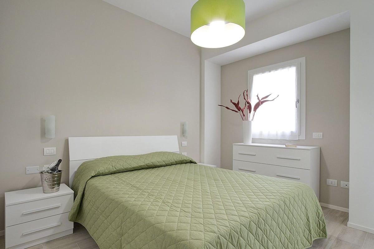 A bedroom with a bed, a nightstand, a dresser, a window, and a green pendant light.
