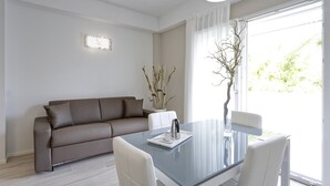 A modern living room with a glass dining table, white chairs, a brown sofa, a vase with branches, and a window with sheer curtains. - Residence Armony Misano (Misano Adriatico)