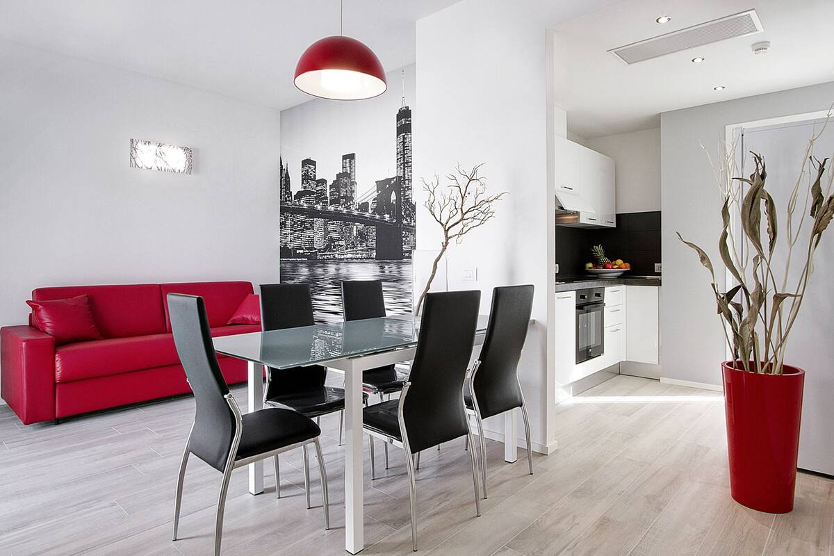 A modern living room with a red sofa, a glass dining table with black chairs, a cityscape wall art, and a red vase with dried plants.