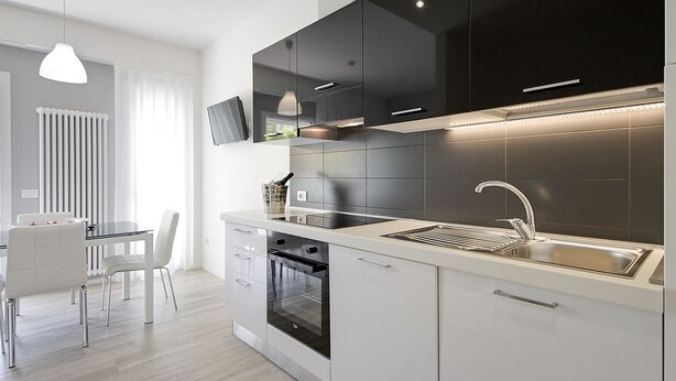 A modern kitchen with a built-in oven, a stainless steel sink, and a dining area with a white table and chairs. - Residence Armony Misano (Misano Adriatico)