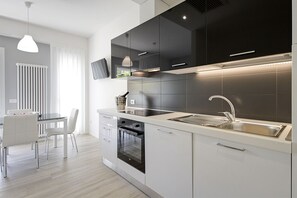 A modern kitchen with a built-in oven, a stainless steel sink, and a dining area with a white table and chairs. - Residence Armony Misano (Misano Adriatico)
