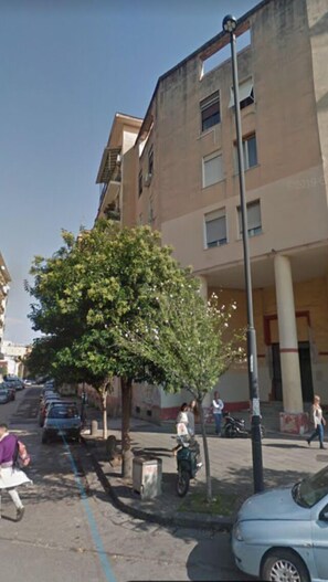 A multi-story building with a tree in front, parked cars, and a few pedestrians. - Voglia di Napoli (Naples)