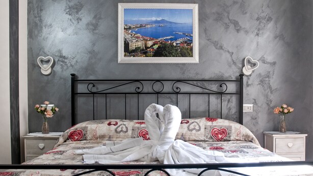 A neatly made bed with a floral bedspread, a heart-shaped towel, and a framed picture of a coastal city on the wall. - Voglia di Napoli (Naples)