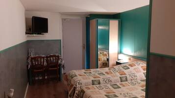 Een kleine, smalle kamer met een bed, een stoel, een tafel en een aan de muur bevestigde televisie.