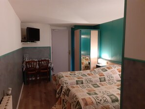 Una habitación pequeña y estrecha con una cama, una silla, una mesa y un televisor fijado a la pared.
