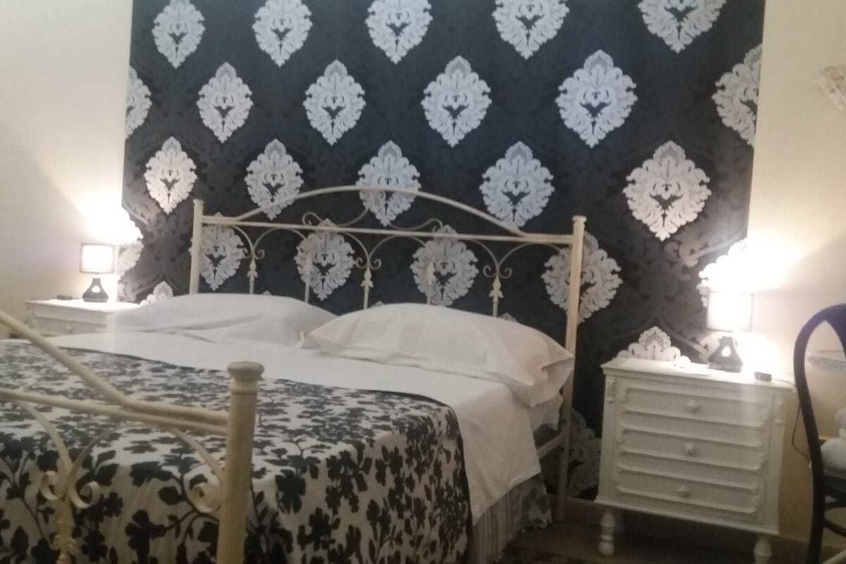 A bed with a metal frame, a bedside table with a lamp, a chair, and a patterned wall.